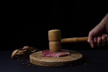 Obraz premium A man's hand beats the meat with a kitchen hammer. Wooden kitchen hammer for meat. Meat on black background