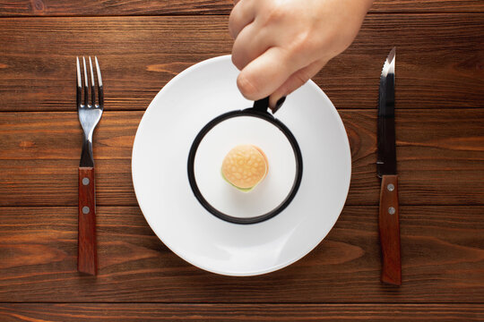 Diet concept. Female hands holding magnifier over little burger on plate. Top view, flat lay.