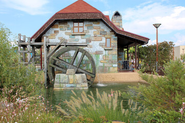 The water mill was built of colored stone under a tiled roof. A wooden wheel, from a gutter, water pours into the pond and rotates the wheel. Flowers and shrubs grow along the shore of the pond.