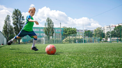 Little boy in blue and green form playing football on open field in the yard, a young soccer player practicing on the football field, moment of hitting the ball