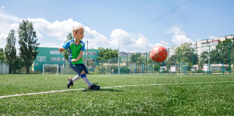 Little boy in blue and green form playing football on open field in the yard, a young soccer player practicing on the football field, moment of hitting the ball