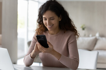 Happy Hispanic young woman sit at desk look at cellphone screen text message online on modern...