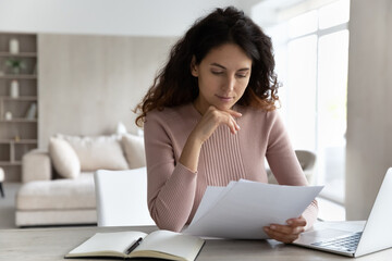 Pensive young Hispanic woman sit at desk at home office work online on computer read paperwork documents. Millennial Latino female use laptop consider financial report or paper correspondence.