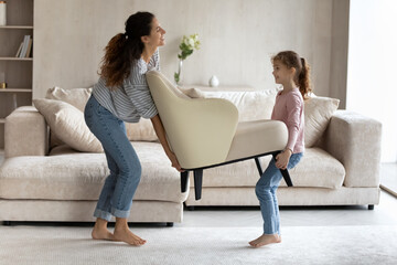 Smiling young single Hispanic mother and teen daughter carry armchair settle in new own home. Happy...