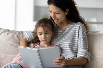 Loving young Latino mother read book to small teen ethnic daughter relax together on couch in...