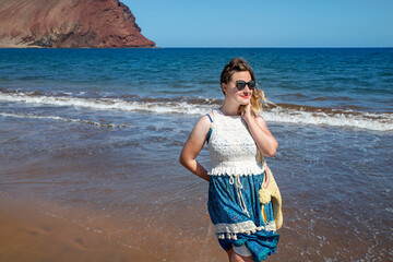 Young millennial woman wearing a blue and white paisley dress, a beach bag and sunglasses, smiling...