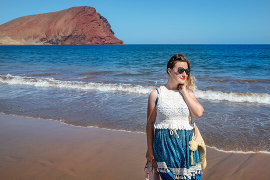 Dreamy holidays portrait of a young millennial woman wearing a blue and white dress and sunglasses while holding her sandals in one hand and walking on Tejita beach in Tenerife, Canary Islands, Spain