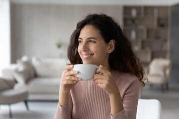 Happy millennial Latino woman drink coffee or tea from mug cup look in distance dreaming thinking of opportunities achievements. Smiling Hispanic female enjoy leisure free time at home visualizing.