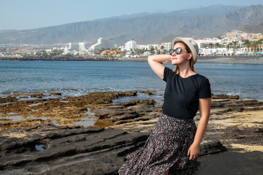 Young millennial woman wearing a hat and sunglasses, enjoying her vacation on the coast at Playa de Las Americas, with the entire Costa Adeje resort in the background, Tenerife, Canary Islands, Spain