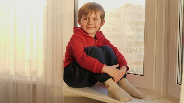 Portrait of smiling little boy sitting on windowsill and looking in camera. COncept of staying at home and covid-19 pandemic
