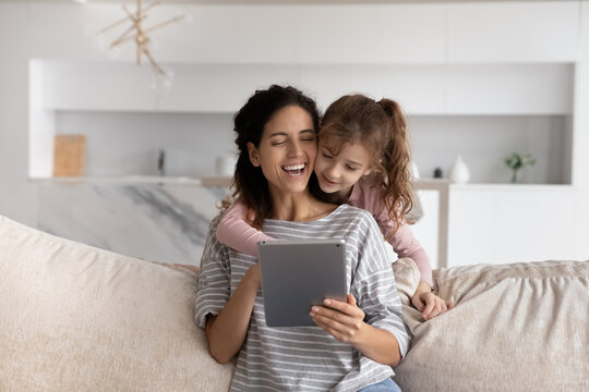 Happy Young Hispanic Mom And Small Teen Daughter Have Fun Use Modern Tablet Gadget At Home Together. Smiling Latino Mother And Little Girl Child Talk Speak On Online Video Webcam Call On Pad Device.