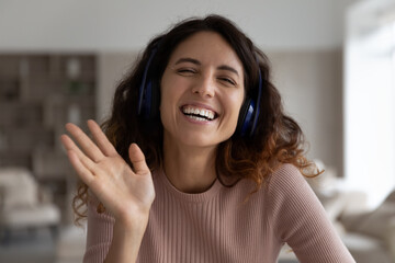 Headshot portrait of smiling millennial Latino female in earphones wave greet talk speak on video...