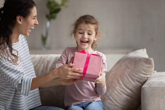 Smiling young Latin mom give wrapped gift box to excited small biracial daughter congratulate with birthday. Happy loving Hispanic mother make surprise present to overjoyed ethnic girl child at home.