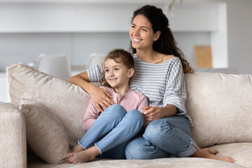 Smiling Hispanic young mother and happy small biracial daughter sit on sofa at home look in distance dream visualize together. Excited Latino mom and little ethnic girl child relax. Vision concept.