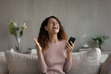 Overjoyed millennial Hispanic woman feel euphoric win online lottery on cellphone gadget. Smiling young Latino female relax at home triumph read good news get promotion notice on smartphone.