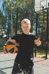 Stylish sportive little kid boy with basketball ball shows thumb up gesture in sunlight. Healthy children lifestyle