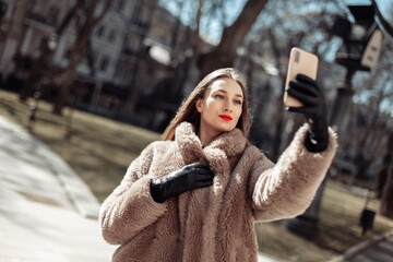 Attractive young fashion woman in a luxurious fur coat makes selfie on smartphone at bright sunny day