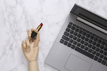 Beauty blog. Wooden hand holds red lipstick and laptop on marble surface. Top view