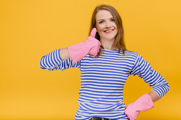 Attractive housewife woman wear rubber cleaning gloves and casual blue striped sweater show thumb up gesture with one hand, body language home cleaning concept standing against a yellow background