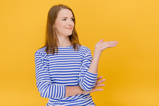 Young Woman Wear Blue Striped Sweater Looking Away Shows Something On Blank Space With Open Hand, Body Language Concept Standing Isolated Against A Yellow Background