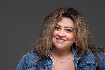Portrait of a beautiful fat woman in a denim jacket looking at the camera and smiling on a gray...