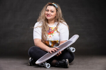 Fototapeta na wymiar Beautiful fat woman with a skateboard on a gray background looks at the camera and smiles.