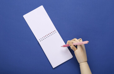 Wooden hand writes with pen in notebook on blue background