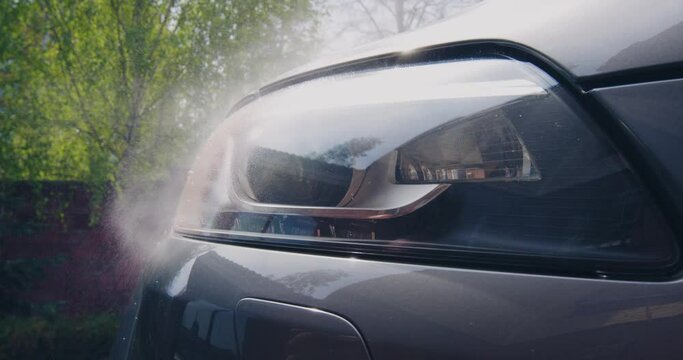 Car detailing concept. Spraying of soapy chemistry for washing headlight on car surface at car wah outdoor. Close up