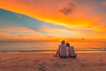 Romantic couple on the beach at colorful sunset on background. Beautiful tropical sunset scenery, romance couple sitting and watching the sunset sea and sky, horizon. Travel, honeymoon destination