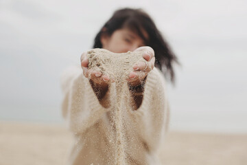 Woman hands holding sand on beach, carefree moment. Stylish young woman in sweater releasing sand on coast. Sand running through hands. Time concept. Summer beach and vacation © sonyachny