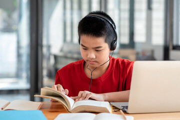 Asian boy wearing headphones while reading a book the laptop  placed on the table in the room.