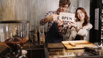 Business owner attractive young Asian woman in apron hanging we're open sign on front door  welcoming clients to new cafe. Happy waitess with protective face mask holding open sign at cafe .