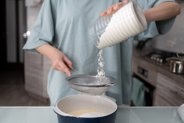 A girl in the kitchen tosses flour from a can into a ceramic bowl for beating eggs with a blender. The concept of making sweet pastries at home.