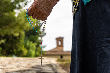 Older woman's hand and arm holding a rosary, near a church. 