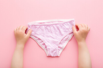 Baby girl hands holding on light purple panties on pink table background. Pastel color. Closeup. Point of view shot. First underwear after diaper using. Top down view. - obrazy, fototapety, plakaty