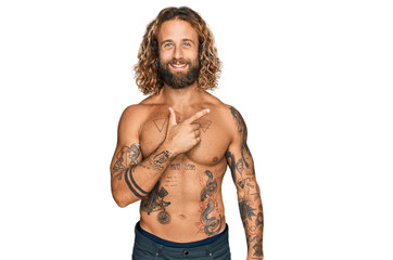 Fototapeta premium Handsome man with beard and long hair standing shirtless showing tattoos cheerful with a smile on face pointing with hand and finger up to the side with happy and natural expression