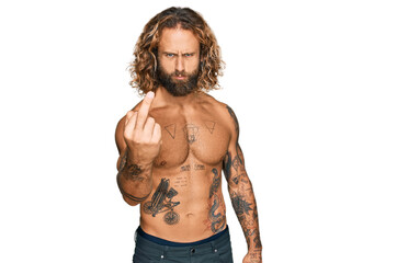 Handsome man with beard and long hair standing shirtless showing tattoos showing middle finger,...