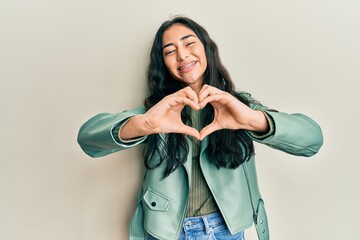 Hispanic teenager girl with dental braces wearing green leather jacket smiling in love doing heart symbol shape with hands. romantic concept. - obrazy, fototapety, plakaty