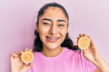 Hispanic teenager girl with dental braces holding lemon smiling with a happy and cool smile on...