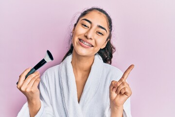 Hispanic teenager girl with dental braces wearing robe holding makeup brush smiling happy pointing...