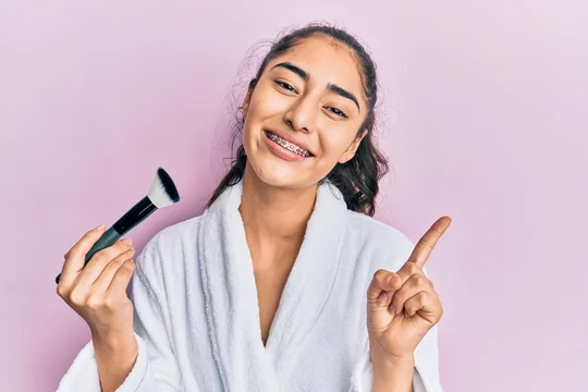 Hispanic teenager girl with dental braces wearing robe holding makeup brush  smiling happy pointing with hand and finger to the side Stock Photo | Adobe  Stock