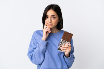 Young caucasian woman isolated on white background taking a chocolate tablet and having doubts