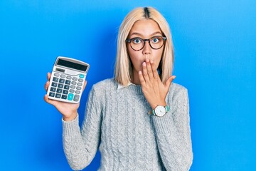 Beautiful blonde woman showing calculator device covering mouth with hand, shocked and afraid for...