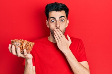 Young hispanic man holding peanuts covering mouth with hand, shocked and afraid for mistake....