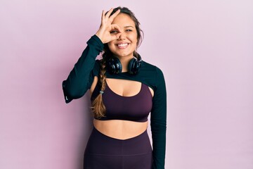 Obraz premium Young hispanic girl wearing sportswear and headphones doing ok gesture with hand smiling, eye looking through fingers with happy face.