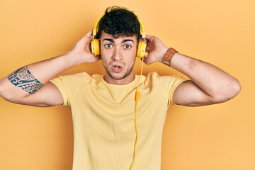 Young hispanic man listening to music using headphones in shock face, looking skeptical and sarcastic, surprised with open mouth