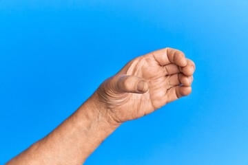 Hand of senior hispanic man over blue isolated background holding invisible object, empty hand doing clipping and grabbing gesture