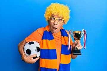 Senior hispanic man football hooligan cheering game holding ball and trophy in shock face, looking skeptical and sarcastic, surprised with open mouth