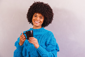 Afro woman dressed for winter, in blue tracksuit, gray background, smiling and warming up, sipping...