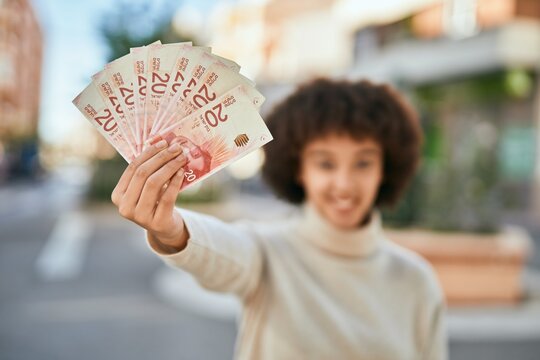 Young hispanic girl smiling happy holding israel shekels at the city.